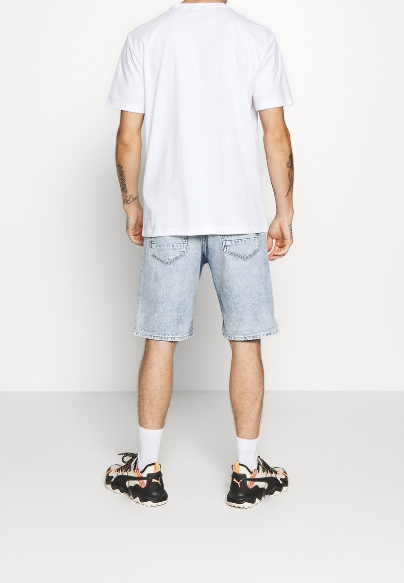Camiseta blanca de manga corta, pantalones cortos de mezclilla azul claro y zapatillas deportivas negras con acentos naranjas. Tatuajes visibles en los brazos.