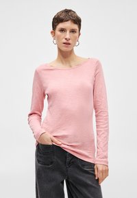 Dlhý rukáv, ľahký, ružový top s dizajnom lodičkového výstrihu a textúrovaným povrchom, nosený s sivými nohavicami, ruka v vrecku.