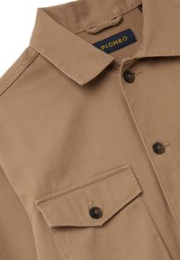 Giacca di camicia color beige realizzata in tessuto liscio, con colletto a punta, chiusura con bottoni e taschino sul petto con patta. Bottoni scuri impreziosiscono il design.