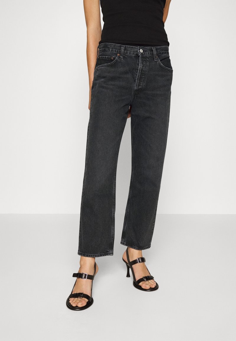 Femme portant un jean noir taille haute à jambe droite accompagné de sandales noires à bout ouvert et d’un haut noir sans manches sur fond blanc.