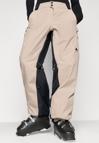 Pantaloni de zăpadă bej și negri, cu buzunare cu fermoar, fermoar la talie și închidere cu nasture. Include manșete elastice și un logo de munte pe lateral.