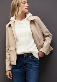 Beżowa kurtka z kołnierzykiem i klapkami, nałożona na białą, futrzaną bluzę z kapturem; ciemnoniebieskie dżinsy. Gładkie tekstury i kontrastujące kolory.