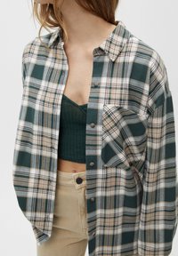 Vrouw in een losvallend groen, beige en witte geruite overhemd met knopen over een donkergroene crop top en beige broek, afgeknipt op borst- en taillehoogte.