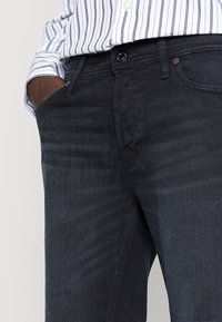 Mörka denimjeans med en mjuk textur och subtila blekningar. Har en knappstängning och standardfickor. Matchas med en randig skjorta.