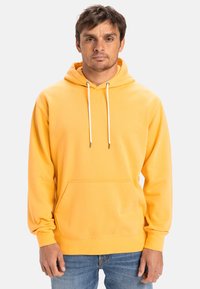 Yellow hoodie with a drawstring hood, front pocket, and embossed logo. Features ribbed cuffs and hem, made from soft fabric. Worn with blue jeans.
