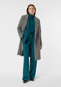 Szary wełniany płaszcz na dopasowanym golfie w kolorze morskim oraz pasujących szerokich spodniach. Czarny pasek podkreśla talię; błyszczące czarne buty dopełniają stylizację.