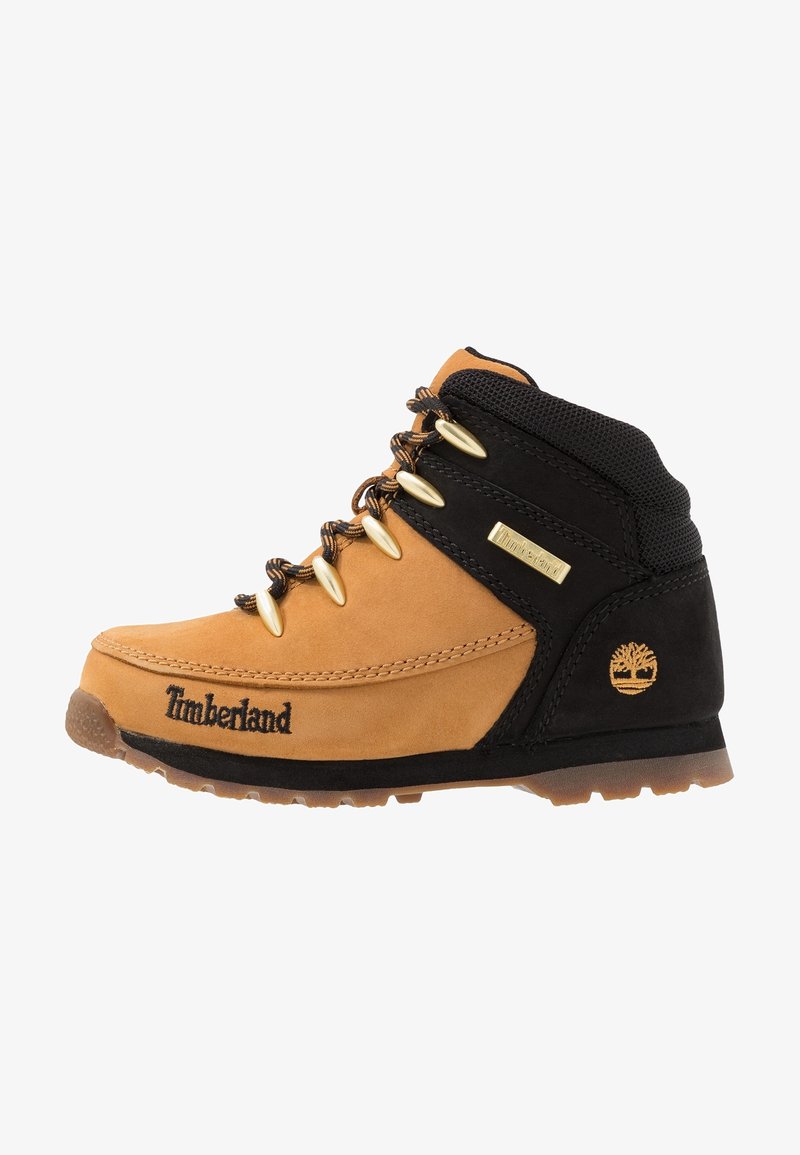 Botte de randonnée Timberland beige et noire avec œillets métalliques, lacets à motifs, semelle en caoutchouc et logo Timberland sur le côté et le talon.