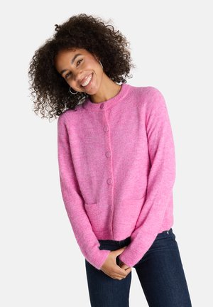 Lächelnde Frau mit lockigem Haar trägt eine rosa knopfgebundene Strickjacke und dunkle Jeans. Sie steht mit gefalteten Händen vor einem weißen Hintergrund.