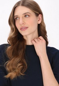 Modella con lunghi capelli castani e mossi, che indossa un top blu navy, illuminata dolcemente, mano sul collo, sfondo neutro. Trucco naturale con dettagli sottili.
