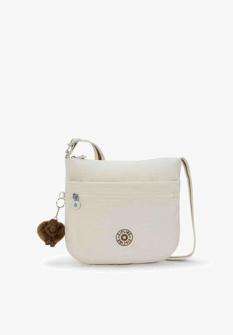 Sac bandoulière beige en tissu doux, doté d'une poche avant zippée, d'une sangle réglable et d'un porte-clés peluche en forme de singe.