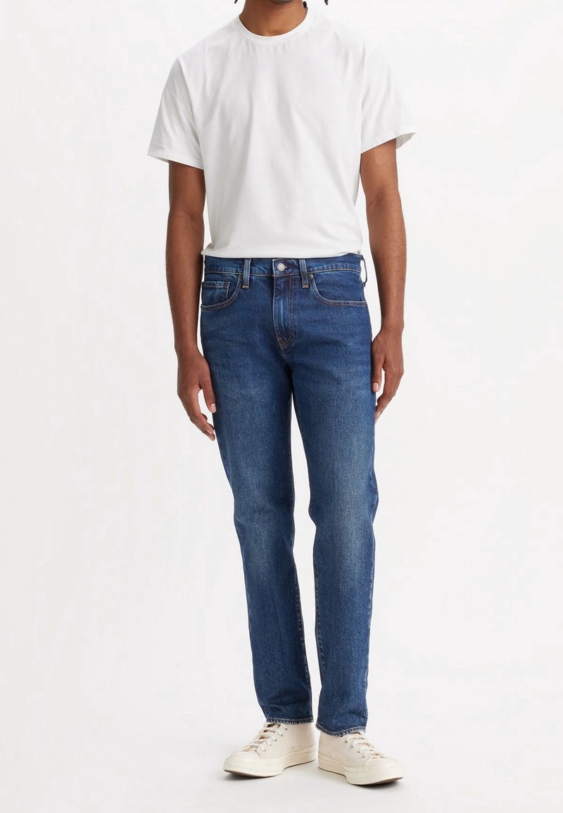 Blue denim jeans with a straight leg fit, paired with a plain white short-sleeve t-shirt and light-colored sneakers.