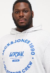 Grau-gestreifte Kapuzenjacke mit einem blauen runden Logo und dem Text "JACK & JONES 1990 COLLECTORS CREW", Kordelzug und strukturiertem Stoff.