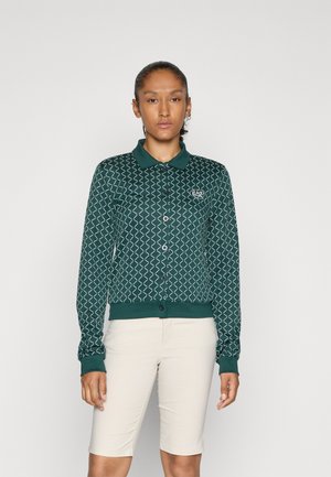 Donna che indossa un cardigan buttonato verde a fantasia con colletto e pantaloni corti beige fino al ginocchio, in piedi contro uno sfondo chiaro e semplice.