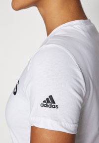 Bílý bavlněný tričko s krátkými rukávy, s černě vyšitým logem Adidas na levém rukávu, hladká textura a standardní střih.