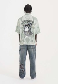 Short-sleeve green patterned shirt featuring an illustrative design on the back, paired with loose-fitting denim pants with embroidered details.