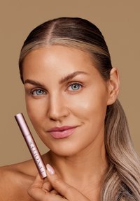 Tubetto di mascara rosa in mano, con design elegante e finitura metallica. La modella ha la pelle liscia, trucco leggero e capelli lunghi e lisci.