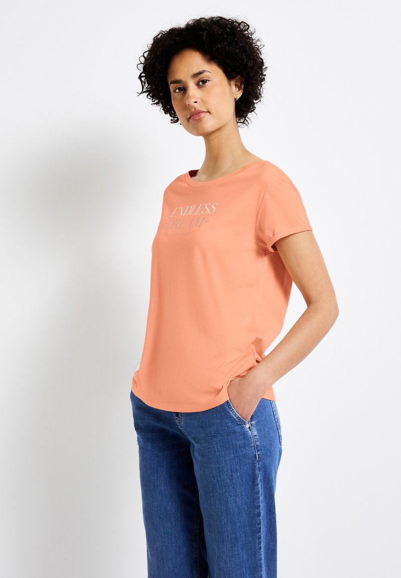 Korálové tričko s krátkým rukávem a kulatým výstřihem, se bílým nápisem "ENDLESS DREAM". V kombinaci s modrými džíny. Hladká textura.
