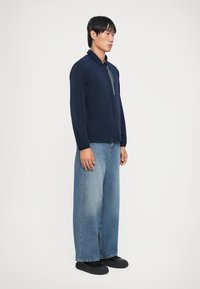 Mornarica zip-up jakna s stojenjem, v kombinaciji z širokimi svetlo modrimi traper hlačami in črnimi čevlji. Gladka tkanina in sproščen kroj.