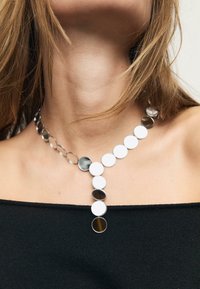 Metalhalskæde med vekslende sorte og hvide cirkulære vedhæng. Designet har en Y-form og skinnende, reflekterende overflader. Accenter i sølv.