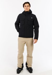 Juodas vandeniui atsparus švarkas su užtrauktuku, dviem krūtinės kišenėmis ir reguliuojama kapuce, derinamas su smėlio spalvos kelnėmis ir juodais slidinėjimo batais.