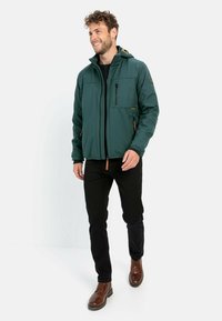 Grøn jakke med hætte, lynlåslukning, flere lommer og orange detaljer. Bæres med sorte bukser og brune støvler. Glat tekstur.