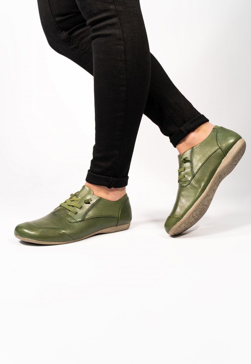 Scarpe in pelle verde con punte rotonde, design con lacci e suole texturizzate, abbinate a jeans neri aderenti su uno sfondo bianco.