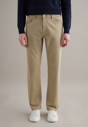 Uomo che indossa pantaloni beige a taglio dritto, maglione blu navy e scarpe da ginnastica bianche, in piedi contro uno sfondo neutro e semplice.