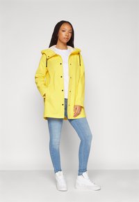 Yellow raincoat with a hood, snap buttons, and drawstrings, worn over a white shirt and blue jeans. Features a relaxed fit and pockets.