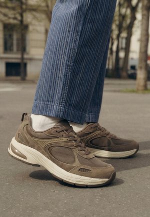 Persoon die bruine mesh- en suède sneakers draagt met witte zolen en witte sokken, staand op het trottoir in blauwe broek met krijtstreep buiten.