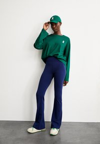 Groene longsleeve shirt met een wit logo, marineblauwe flared broek en beige sneakers met groene accenten. Eenvoudige, casual sportoutfit.