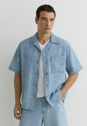 Bărbat purtând cămașă cu mânecă scurtă, albastru deschis cu model, peste un tricou alb și blugi albastru deschis, stând cu o mână în buzunar.