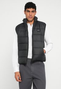 Melnā pūkstošā veste ar augstu apkakli, izšūtu dizainu un priekšējo rāvējslēdzēju. Valkāta virs baltas garām piedurknēm krekliņa, tajā ir sānu rāvējslēdzēja kabatas.