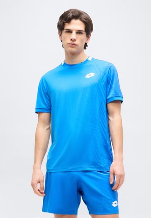 Νεαρός άνδρας με κοντά καστανά μαλλιά που φοράει φωτεινό μπλε αθλητικό πουκάμισο και αντίστοιχα σορτς, σε απλό φωτεινό φόντο.