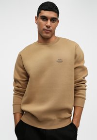 Fiatal fiú rövid göndör hajjal, teveszínű pulóverben, melynek mellén kicsi fekete logó felirat található, kezei feketében nadrág zsebében.