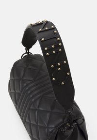 Black quilted leather handbag with a thick strap featuring gold studs and an embossed logo. The design includes a side zipper and textured accents.