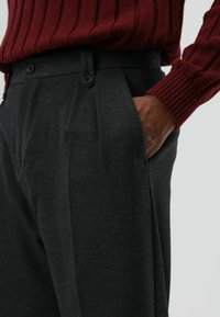 Op maat gemaakte grijze geplooide broek met een gladde textuur, voorzien van twee voorzakken en zwarte knopen bij de tailleband, gecombineerd met een geribbelde bordeauxrode trui.