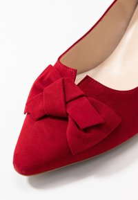 Bottine en daim rouge avec un avant pointu et un grand nœud à l'avant. Intérieur lisse et coutures subtiles le long des bords.