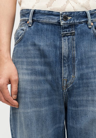 Μπλε τζιν παντελόνι με ξεθωριασμένη υφή, κλείσιμο με κουμπιά και πορτοκαλί ραφές. Κοντινή λήψη, χέρι ακουμπισμένο στο ύφασμα, υπογραμμίζοντας το σχέδιο.