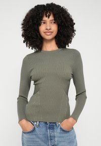 Φιλικό μακρυμάνικο τοπ με ραβδώσεις σε ελιά, με ανάγλυφα πάνελ και στρογγυλή λαιμόκοψη, φορεμένο με ανοιχτό μπλε τζιν παντελόνι.