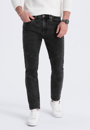 Mann trägt schwarze Slim-Fit-Jeans mit Nahtdetails, weiße Sneaker, einen weißen Pullover, eine schwarze Uhr und ein Armband, steht mit den Händen nahe den Taschen.