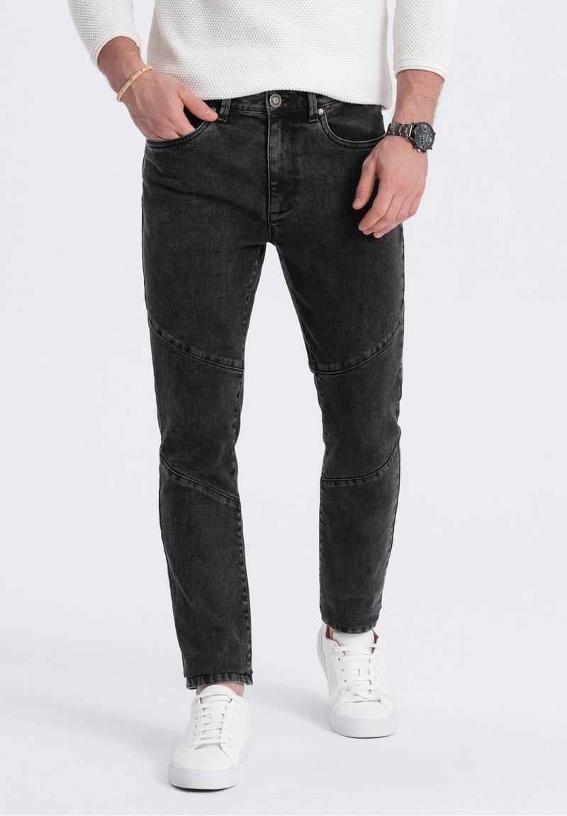 Homme portant un jean slim noir avec des détails de couture, des baskets blanches, un pull blanc, une montre noire et un bracelet, debout les mains près des poches.
