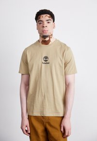 Bežinis medvilninis marškinėlis su juodu Timberland logo ir medžio grafika ant krūtinės, derinamas su rudomis kelnėmis. Paprastas, kasdienis dizainas.