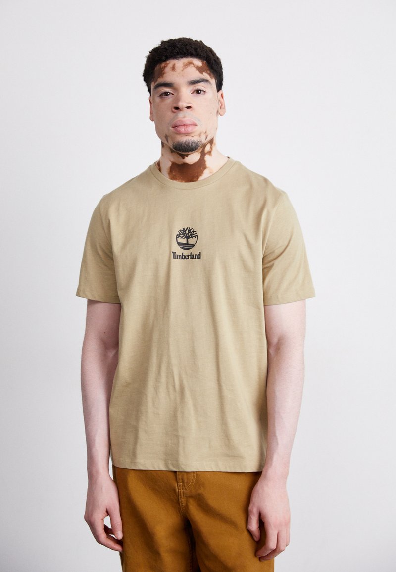 Bežinis medvilninis marškinėlis su juodu Timberland logo ir medžio grafika ant krūtinės, derinamas su rudomis kelnėmis. Paprastas, kasdienis dizainas.