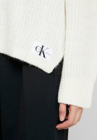 Maglione bianco a maglia con orlo a coste, morbida texture e patch con logo Calvin Klein Jeans sul lato sinistro. Pantaloni scuri parzialmente visibili.