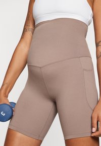 Høyt liv brun sportshorts med glatt tekstur, sidelomme og figursydd design, kombinert med en hvit topp og en blå manual.