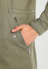 Veste vert olive avec une poche latérale zippée, une texture de tissu douce et une fermeture éclair en métal contrastée. Design simple et épuré.