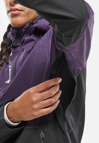 Lila und schwarze Jacke mit Reißverschlüssen, ausgestattet mit hohem Kragen, verstellbare Kapuze und strukturiertem Stoff. Eine Hand greift in eine Seitenasche.