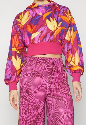 Kobieta nosząca kolorowy krótki hoodie w żółto-pomarańczowy kwiatowy wzór oraz różowe spodnie w wysokim stanie z wzorem.