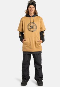 Beżowa bluza z kapturem z dużym czarnym logo z przodu, w połączeniu z czarnymi spodniami śnieżnymi i czarnymi butami snowboardowymi. Gogle narciarskie na głowie.