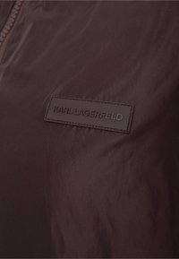 Close-up of dark fabric with a stitched rectangular label embossed with "KARL LAGERFELD" near a zipper.